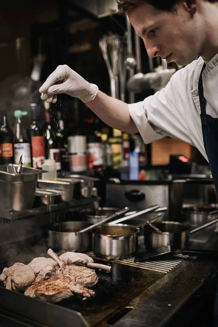 Chef's hand seasoning meat on the grill in a busy kitchen.